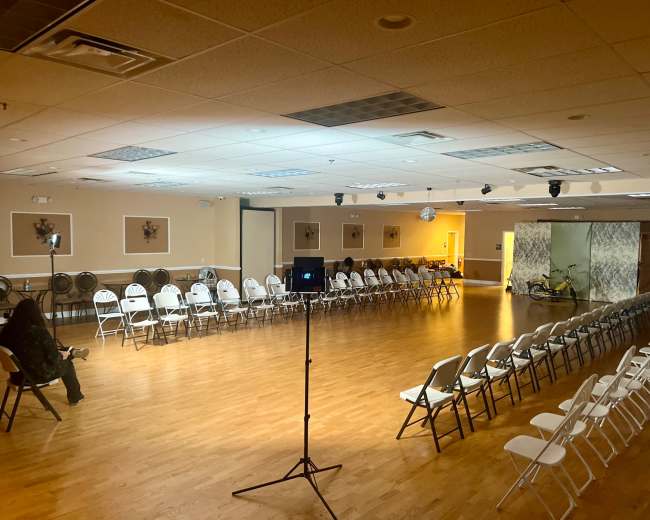 A spacious event room with rows of white chairs arranged in two parallel lines, and a small stage set at one end.