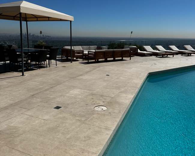 A clear blue pool is surrounded by lounge chairs and a dining area with a canopy on a sunlit terrace overlooking a cityscape.