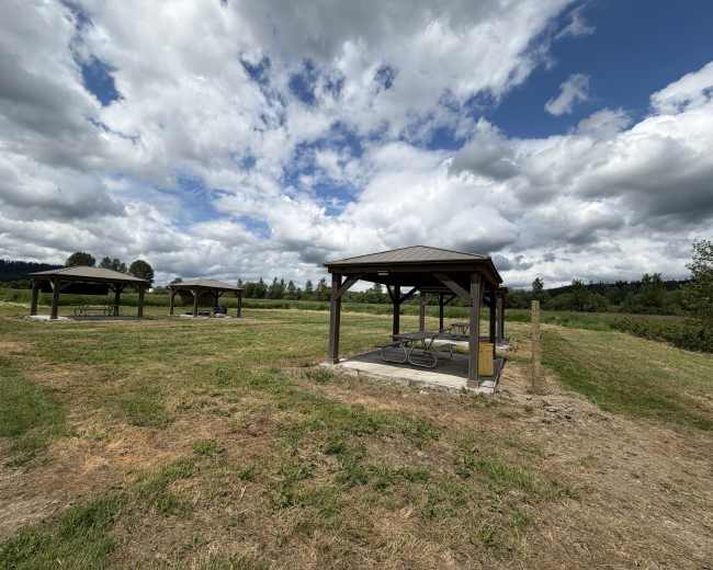 The image shows several wooden picnic shelters on a grassy field under a partly cloudy sky.