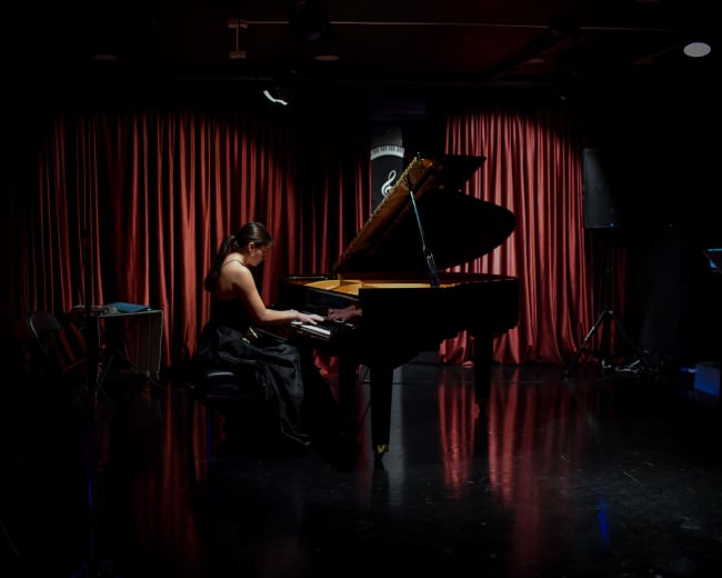 A woman in a black dress plays a grand piano on a dimly lit stage with red curtains.
