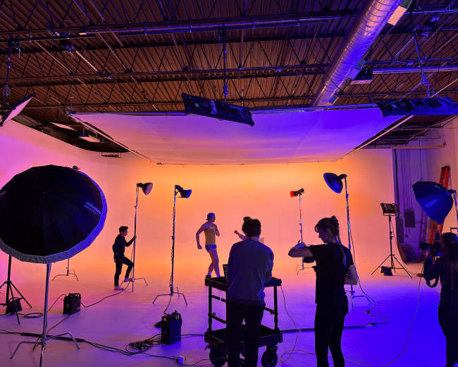 A studio space is set up for a photoshoot, featuring lighting equipment, a backdrop, and several individuals posing and adjusting settings.
