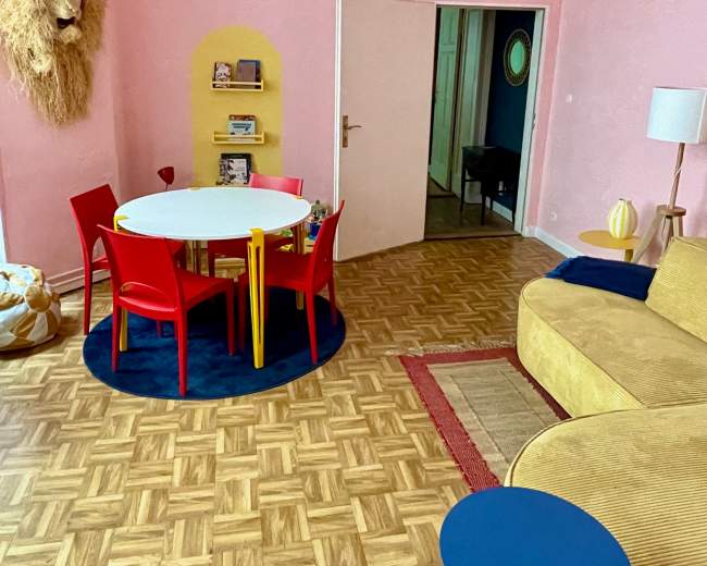 The image shows a colorful living room with a round white table surrounded by red and yellow chairs, a large yellow wall shelf, and a cozy yellow couch against pink walls.