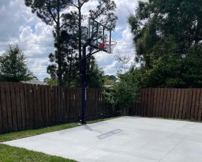 The image shows a concrete basketball court with a hoop and a nearby grassy area and pathway.