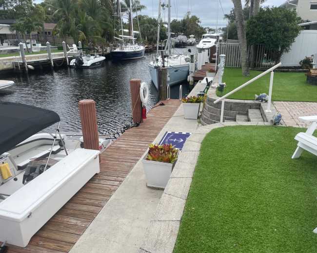 A wooden dock lined with boats leads to a grassy area with decorative planters and steps beside a calm waterfront.