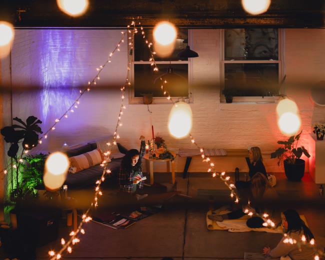 A dimly lit indoor space with string lights, featuring people seated on the floor and engaging in an activity among plants and furniture.