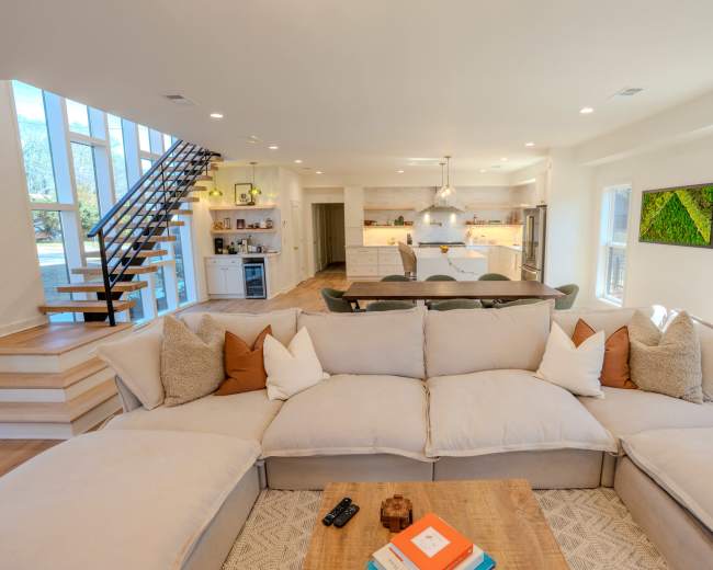A spacious living room features a large sectional sofa with decorative pillows, facing a coffee table, while a staircase to the left leads to an upper level and the kitchen area is visible in the background.