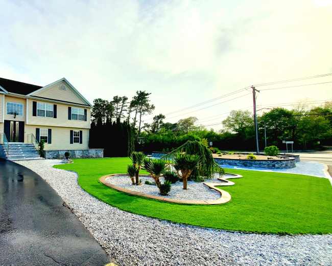 The image shows a two-story house with a garage, surrounded by a well-maintained lawn and landscaping, including gravel paths and small palm trees.