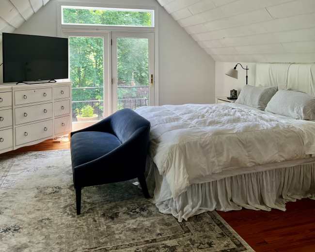 The image shows a bright, airy bedroom with a large bed, a dark blue chair, a dresser, and windows overlooking a wooded area.