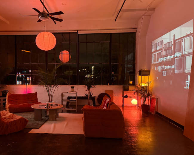 The scene shows a modern, low-light lounge with bright orange furniture, multiple retro televisions, and a large wall projection, creating a cozy ambiance.
