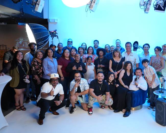A group of approximately 40 people poses together in a studio with a blue backdrop and professional lighting equipment.