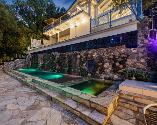 A modern house with a stone-accented pool features colorful lighting and a waterfall, set in a landscaped outdoor area.