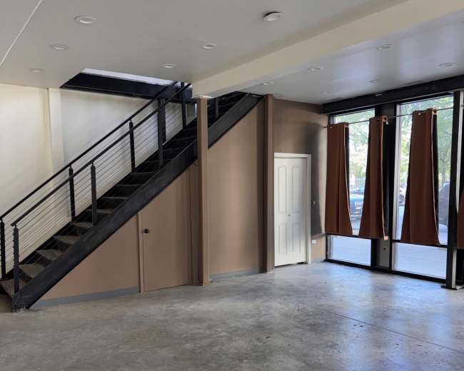 The image shows an empty room with concrete flooring, a staircase on the left, and large windows covered with brown curtains.