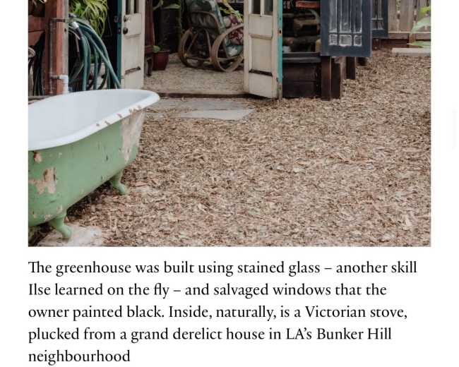 The image shows a greenhouse constructed with stained glass and salvaged black-painted windows, set in a garden area featuring a Victorian stove.