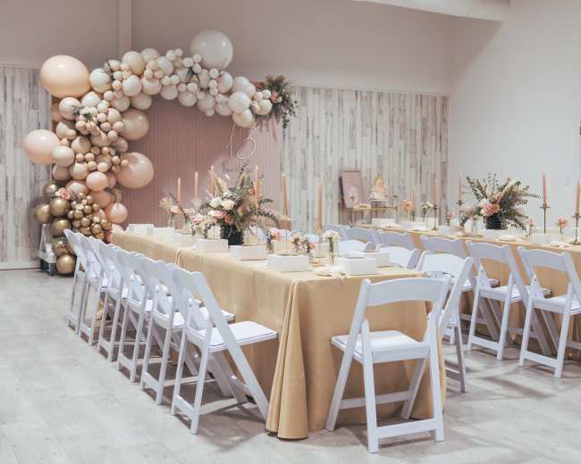 A beautifully arranged event space features a long table set for a gathering, surrounded by floral decorations and an arch made of balloons.