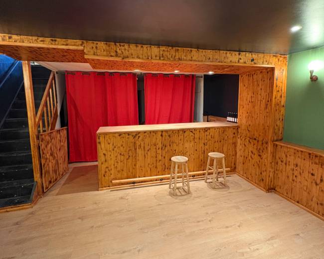 The image shows a basement bar area with wooden walls, a red curtain backdrop, and two stools in front of a counter.