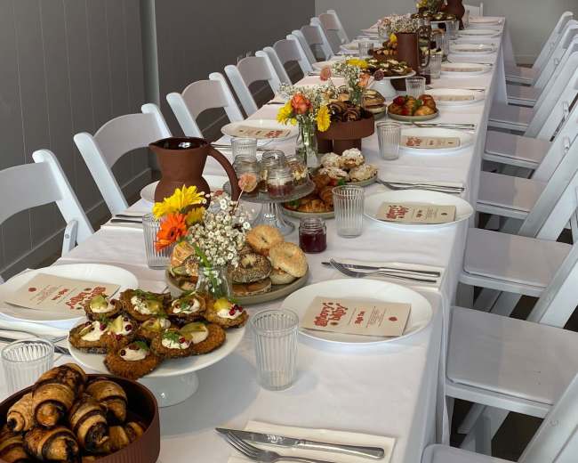 A long dining table is set with various dishes and decorated with flowers, under a neon sign that reads "Baba."