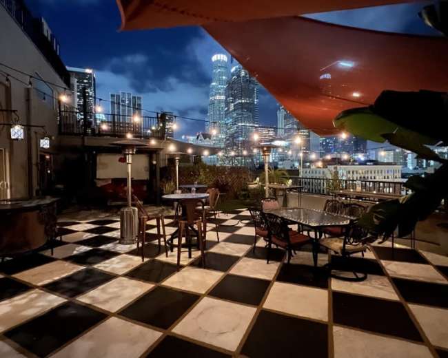 A rooftop patio with black and white checkered tiles, surrounded by city buildings and illuminated by string lights and a nearby structure.