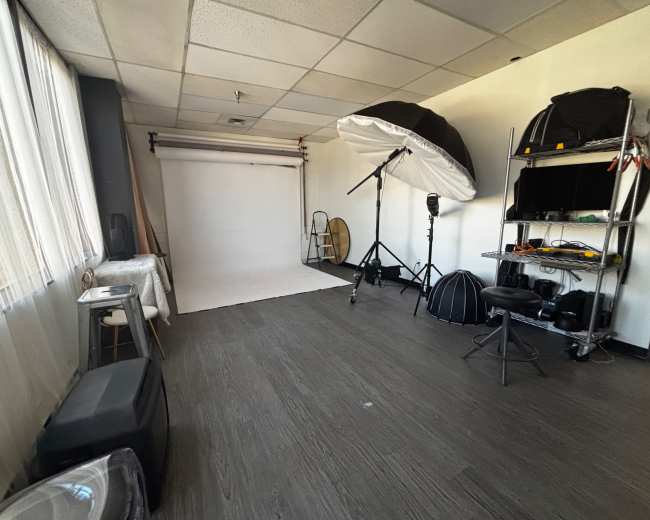 The image shows a photography studio with a white backdrop, various lighting equipment, and props arranged along the walls.