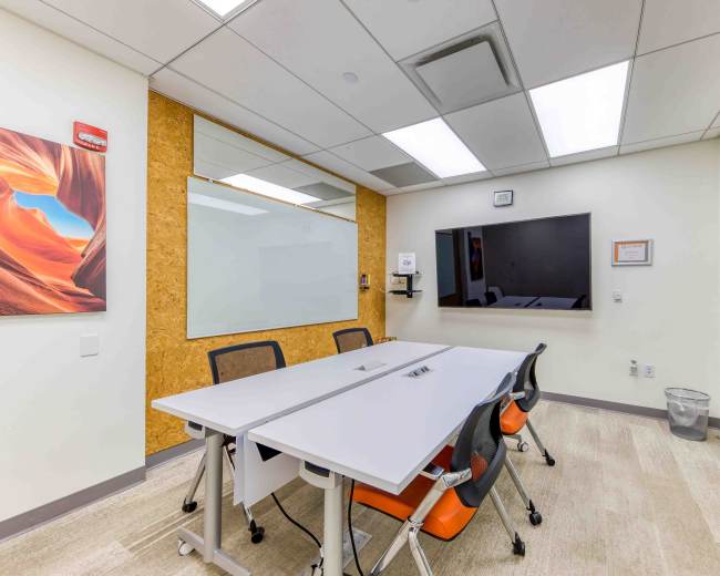 A conference room features a white table surrounded by orange and gray chairs, a wall-mounted screen, and a whiteboard.