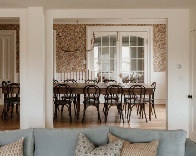 A long wooden dining table surrounded by chairs is visible through an open doorway, with patterned wallpaper and large glass doors in the background.