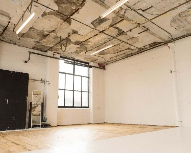 The image shows an empty industrial-style room with wooden flooring, a large window, and exposed ceiling elements.