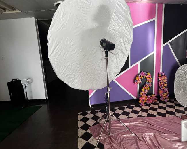 A large white umbrella reflector stands on a tripod in a room adorned with colorful decorations and geometric wall designs.