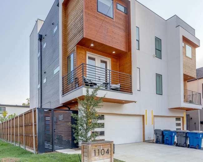 The image shows a modern two-story building with a combination of wooden and gray siding, featuring balconies and a parking area with blue trash bins.