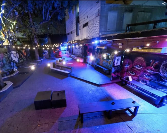 An outdoor bar area with string lights illuminating colorful murals on the walls and wooden seating arrangements.