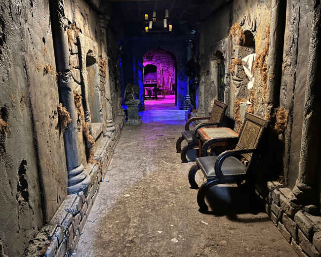 The image shows a dimly lit corridor with textured stone walls, featuring two wooden chairs and an entrance leading to a room illuminated in purple light.