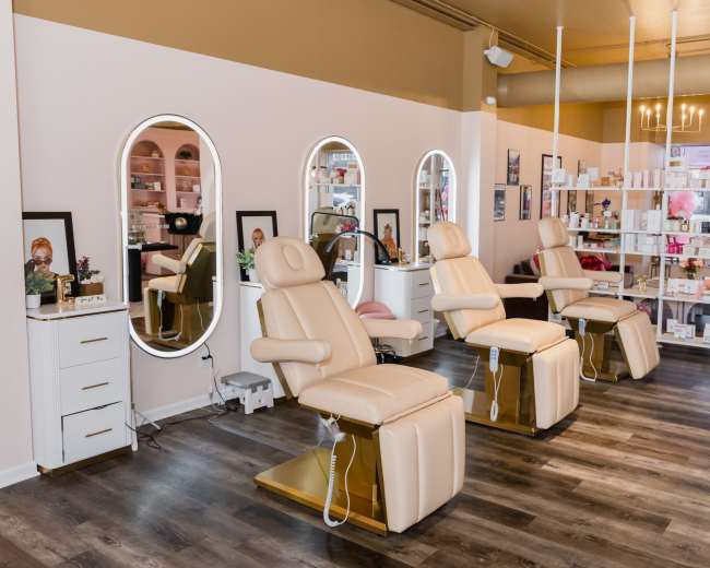 A modern beauty salon with three beige treatment chairs arranged in front of illuminated mirrors against a backdrop of shelves filled with assorted beauty products.