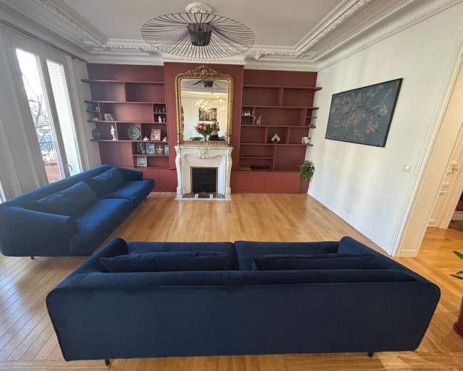 The room features two blue sofas arranged around a white fireplace, with a red bookshelf and decorative items lining the walls.