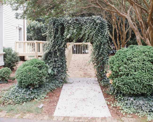 A garden path leading through an arch covered with climbing greenery, flanked by neatly trimmed bushes.