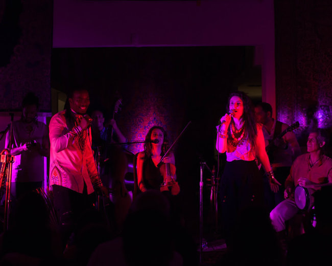 A diverse group of musicians performs on stage with colorful lights illuminating the scene.