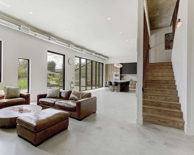 The image shows a spacious, modern living room featuring leather sofas, large windows allowing natural light, and a staircase leading to an upper level.