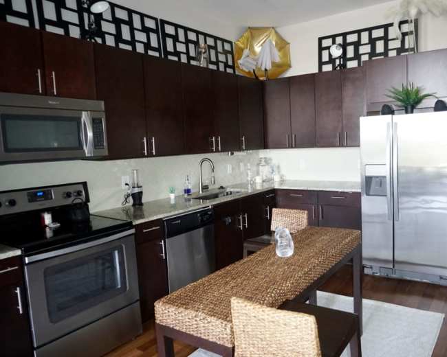 The image shows a modern kitchen with dark wooden cabinets, stainless steel appliances, and a small dining table set with wicker chairs.