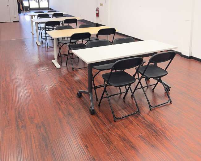 The image shows an empty room with several rows of tables and chairs set up along the wooden floor, illuminated by overhead lights.