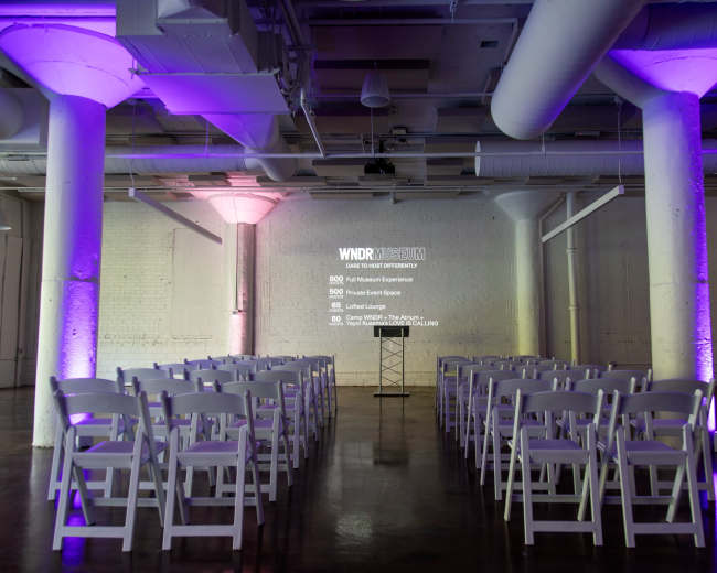 The image shows an empty event space set up with rows of white chairs facing a podium, illuminated by colorful lights on the walls.