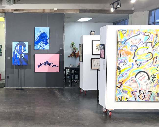 The image shows an art gallery featuring various artworks displayed on white partitions and walls, with a mix of paintings and framed prints.