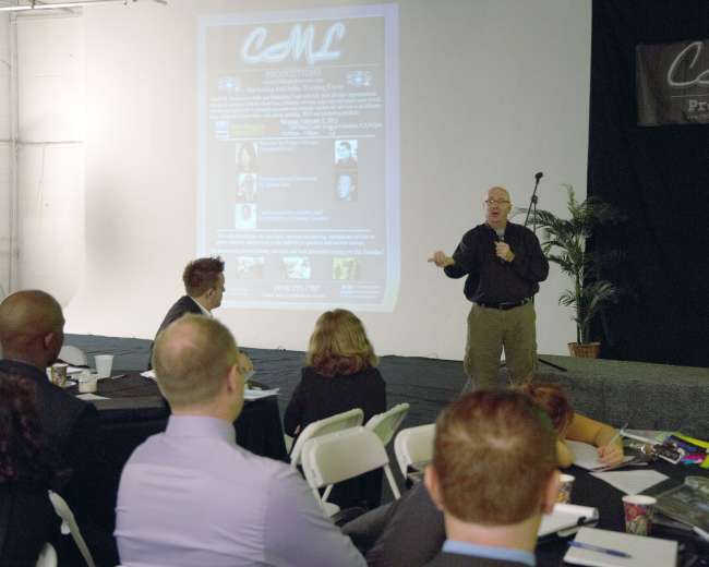 A speaker presents to an audience in a conference room, with a large projection screen displaying information about a production company in the background.