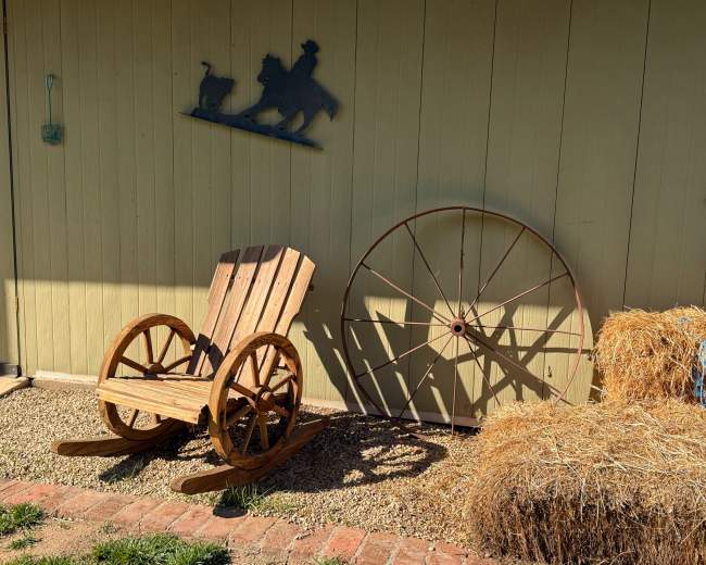 A wooden rocking chair is positioned beside a large wooden wheel and hay bales against a green wall.