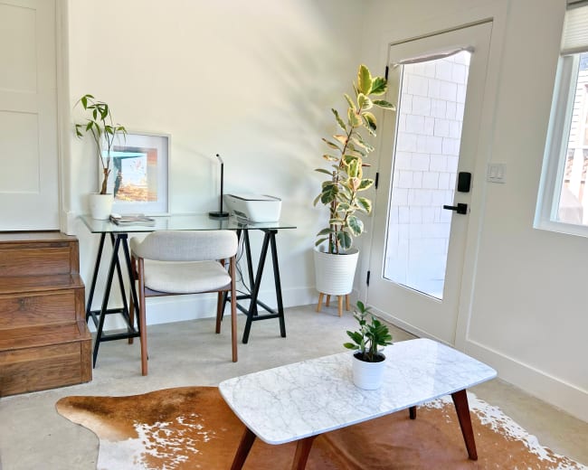 The image depicts a modern home office with a glass desk, a chair, a plant, and a marble coffee table on a cowhide rug near a doorway.