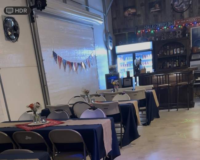 The image shows a spacious venue with rows of tables covered in dark tablecloths, decorated with small vases of flowers, and a bar area in the background.