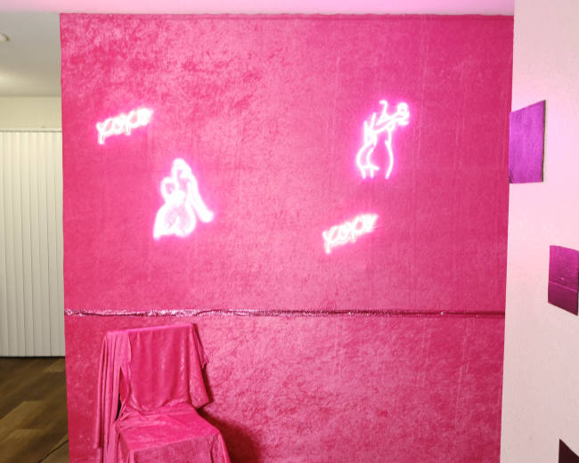 A pink textured wall featuring three neon signs and a covered chair in front of it.