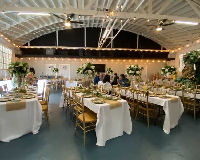 A banquet hall setup with elegantly arranged tables, each featuring gold accents and floral centerpieces, while guests are seated and conversing.