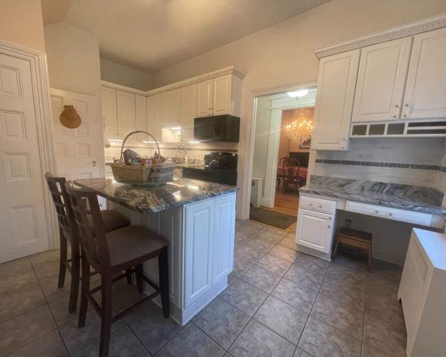 A modern kitchen features white cabinetry, a marble countertop island with seating, and multiple doorways leading to other areas of the home.
