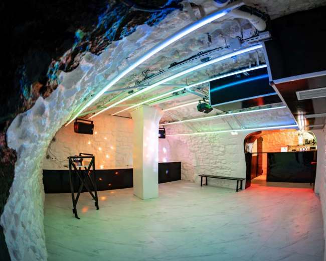 The image shows a modern underground room with stone walls, illuminated by colorful LED lights and features a bar area.