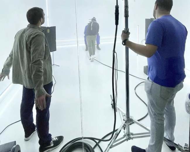 Two crew members are positioned in a brightly lit studio, while a third person walks away into a light-filled area.