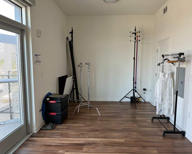 The image shows a sparsely furnished room with wooden flooring, featuring clothing racks, lighting stands, and a large window.