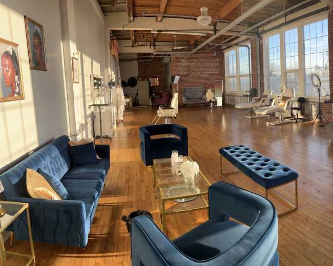 The image shows a spacious, brightly lit room with wooden floors and large windows, furnished with various sofas, chairs, and a coffee table, creating a welcoming atmosphere.