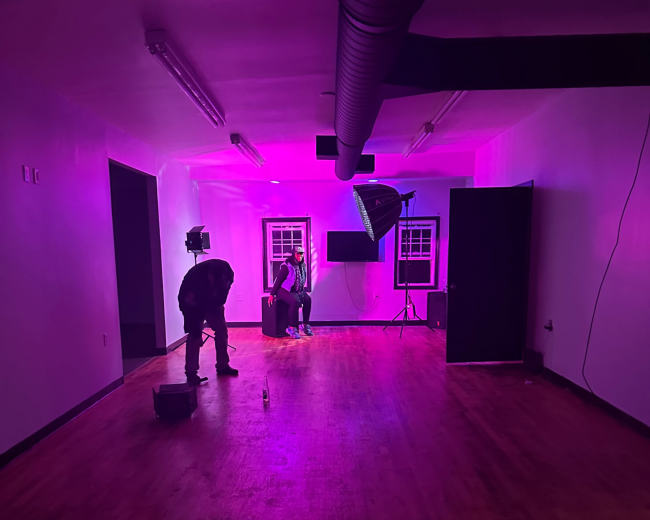 A photographer sets up lighting equipment in a sparsely furnished room with purple illumination while a subject sits on a stool.